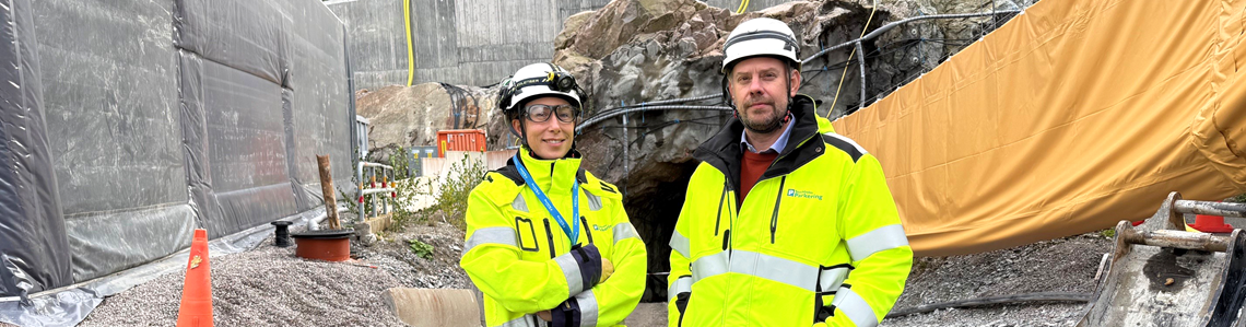 Vd samt projektchef i varselkläder och bygghjälm framför Hjorthagsberget